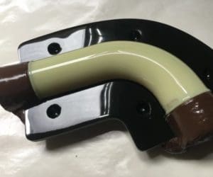 A curved, cream-colored pipe is mounted on a black, glossy surface with four holes. The ends of the pipe are wrapped in brown material.