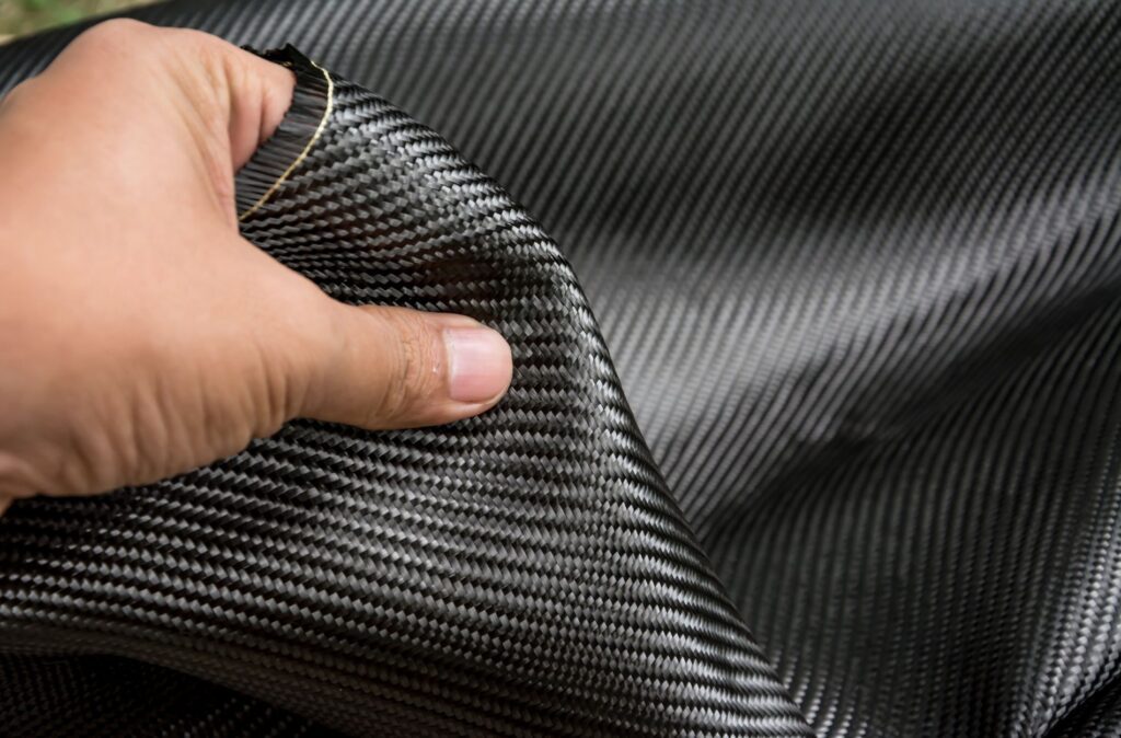 A hand gently grasps a sleek sheet of black carbon fiber fabric, showcasing its intricate woven texture and remarkable flexibility.